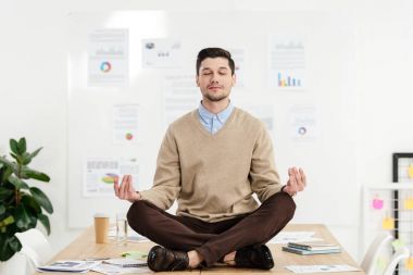relaxed businessman with eyes closed sitting in lotus position on table in office clipart