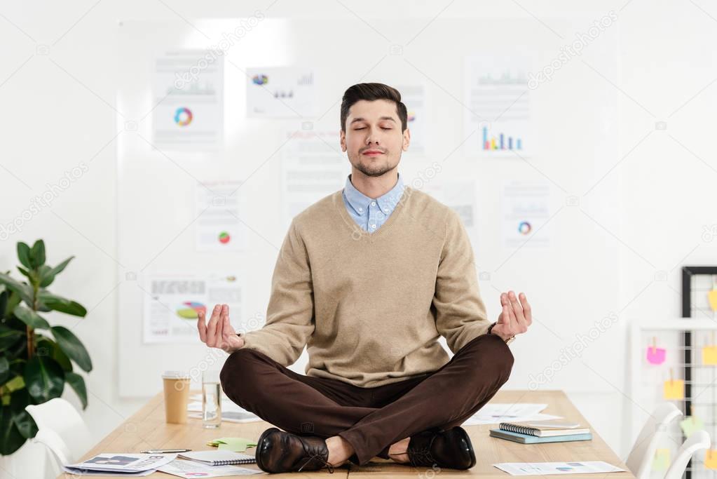 relaxed businessman with eyes closed sitting in lotus position on table in office