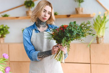 attractive young tattooed woman in apron holding beautiful bouquet in flower shop clipart