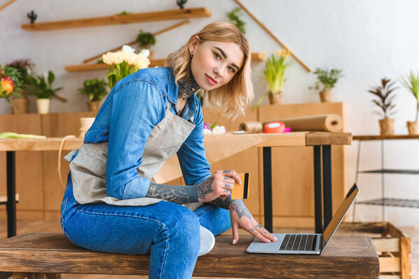 beautiful young florist holding credit card and looking at camera while using laptop in flower shop 