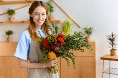 beautiful young florist holding bouquet and smiling at camera in flower shop clipart