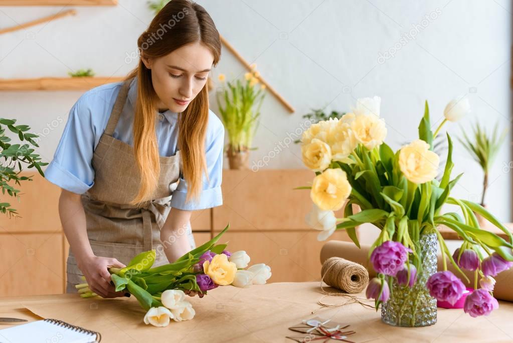 young florist in apron arranging beautiful tulip flowers at workplace