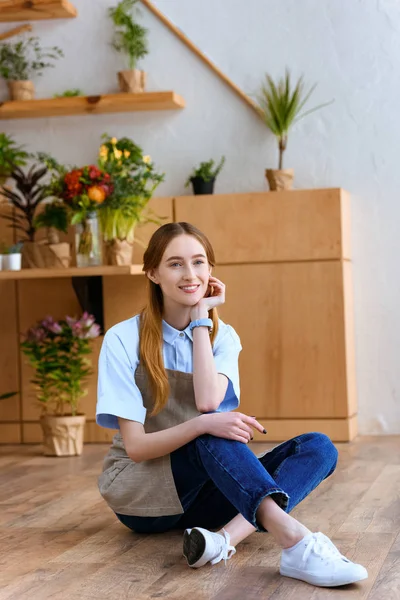 Красива Молода Жінка Фартусі Сидить Підлозі Посміхається Камеру Квітковому Магазині — Безкоштовне стокове фото