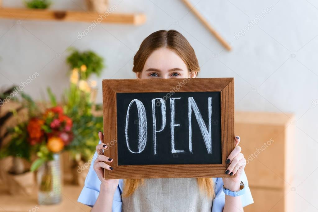 young female florist holding open sign and looking at camera in flower shop