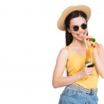 Smiling girl in sunglasses and hat drinking cocktail, isolated on white