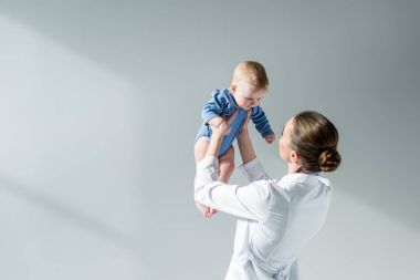 rear view of female pediatrician playing with little baby on grey clipart