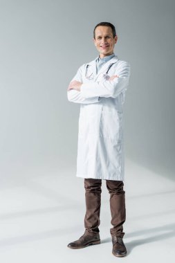 handsome adult doctor with crossed arms looking at camera isolated on grey clipart