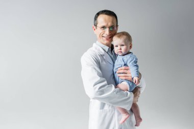 küçük bebek tutan ve gri izole kamera bakarak yakışıklı yetişkin çocuk doktoru