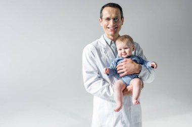 küçük bebek tutan ve gri izole kamera bakarak mutlu yetişkin çocuk doktoru