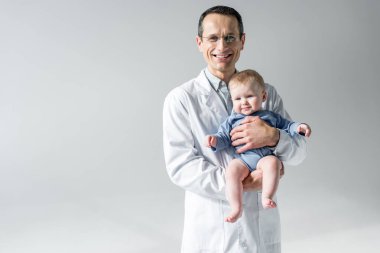 happy adult pediatrician holding little baby and looking at camera isolated on grey clipart