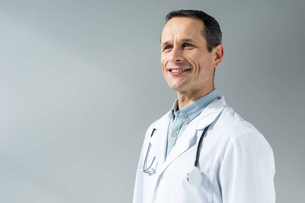 handsome adult doctor in coat looking away isolated on grey