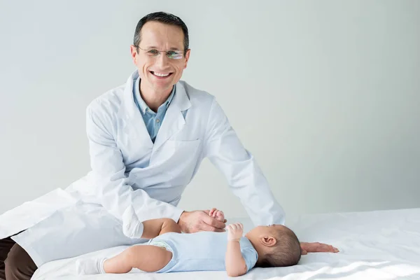 小さな赤ちゃんをベッドの上に座って幸せアダルト小児科医 — ストック写真