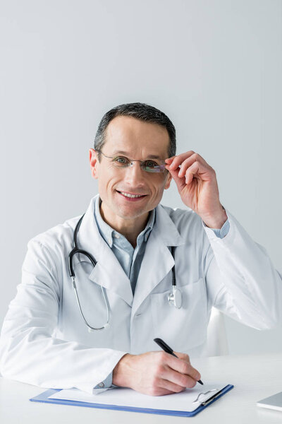 happy adult doctor sitting at workplace and looking at camera while writing in clipboard isolated on white