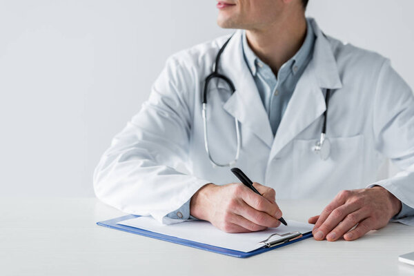 cropped shot of doctor sitting at workplace and writing diagnosis in clipboard isolated on white