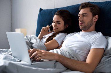 Young man and woman using digital devices while lying in bed clipart