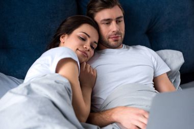 Smiling couple looking at laptop while lying in bed clipart