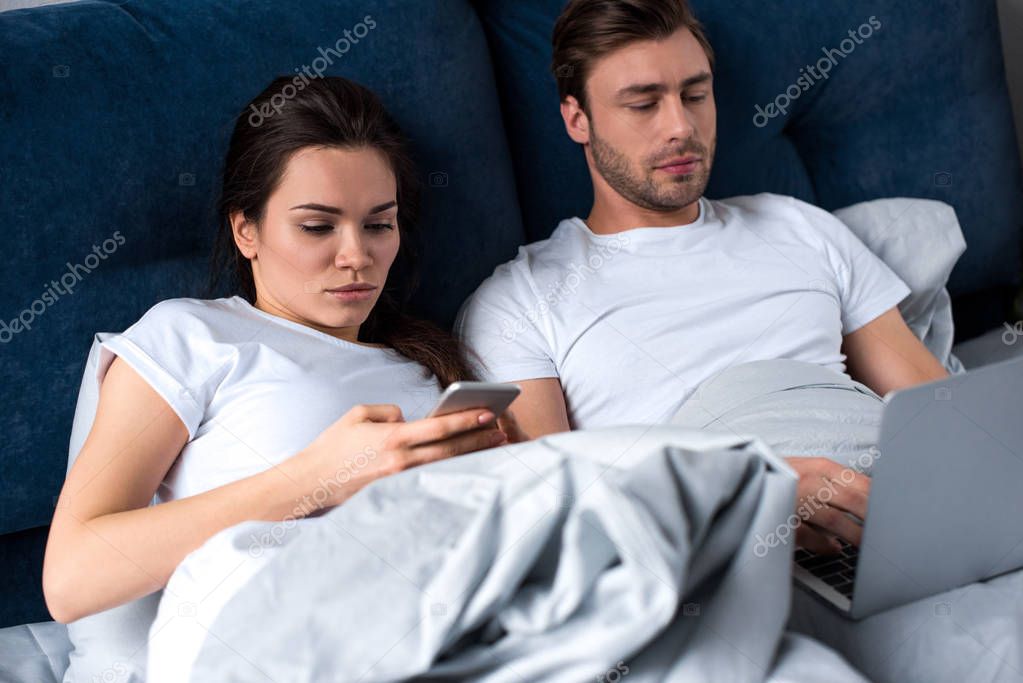 Young man and woman using digital devices while lying in bed