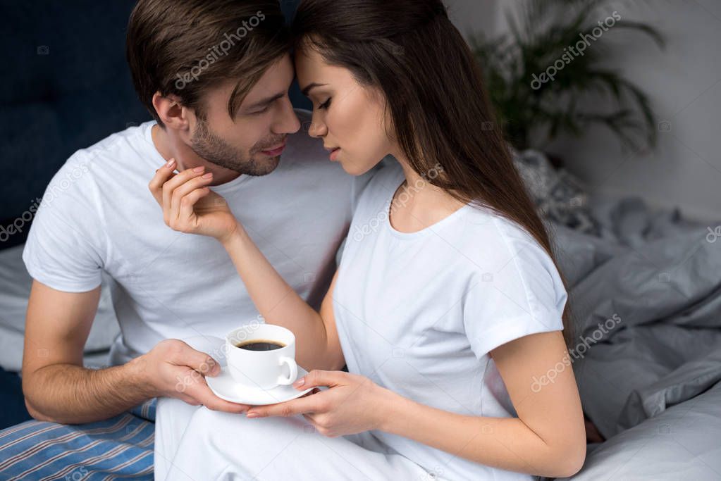 Woman holding cup of coffee and embracing her husband while in bed