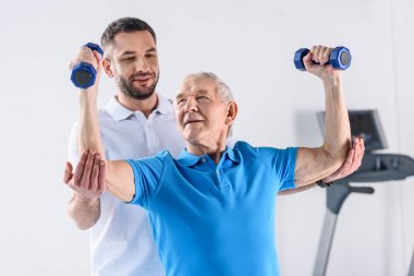 portrait of rehabilitation therapist assisting senior man exercising with dumbbells on grey backdrop clipart