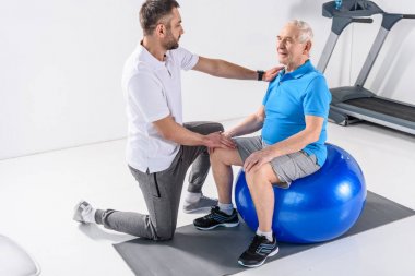 rehabilitation therapist assisting senior man exercising on fitness ball clipart
