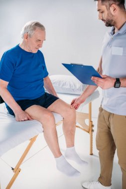 Rehabilitasyon terapist kıdemli kontrol Not Defteri ile diz masaj masasında gri zemin üzerinde mans