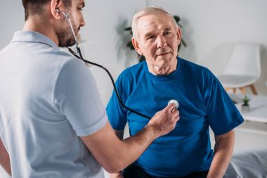 rehabilitation therapist with stethoscope checking senior mans heartbeat