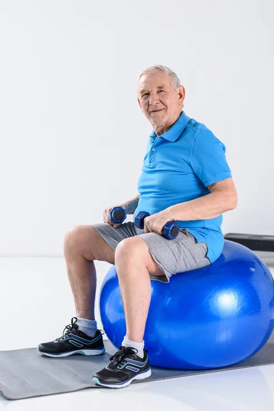 在灰色背景下的健身球上坐着哑铃的微笑老人 — 图库照片