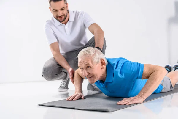 Terapeuta Riabilitativo Che Aiuta Uomo Anziano Esercitarsi Tappeto Sfondo Grigio — Foto Stock