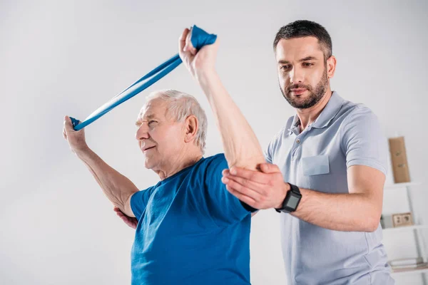 Visão Lateral Terapeuta Reabilitação Ajudando Homem Idoso Exercitar Com Fita — Fotografia de Stock