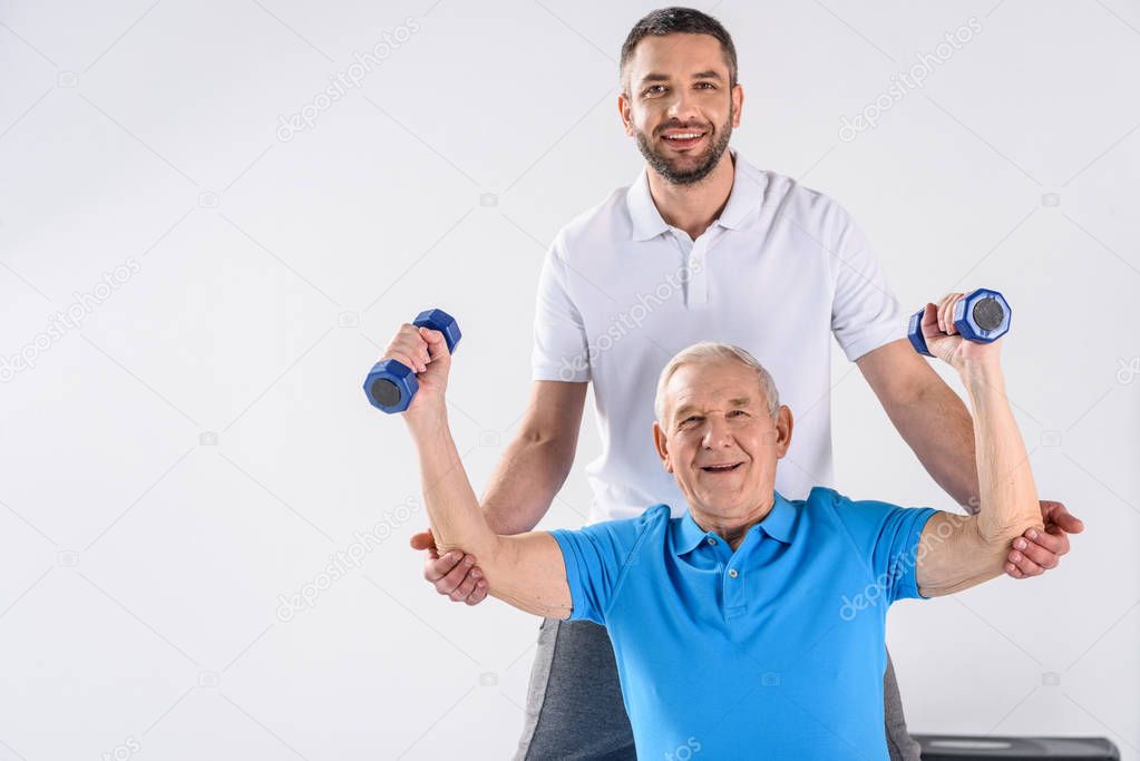 portrait of smiling rehabilitation therapist assisting senior man exercising with dumbbells isolated on grey