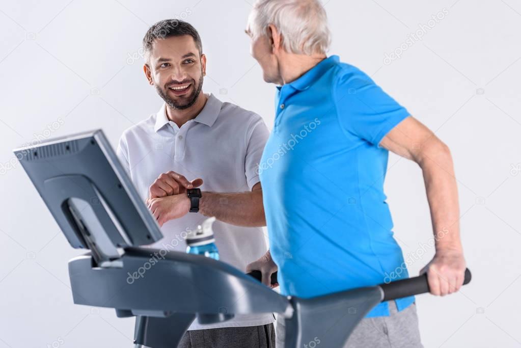 smiling rehabilitation therapist assisting senior man exercising on treadmill isolated on grey