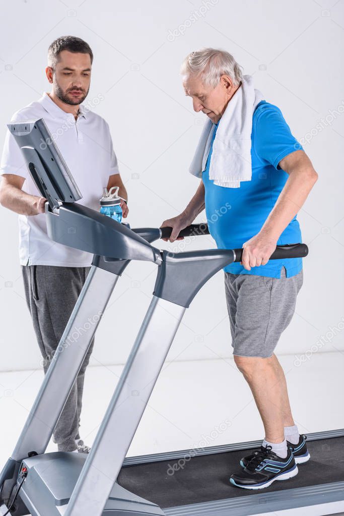 rehabilitation therapist assisting senior man with towel exercising on treadmill isolated on grey