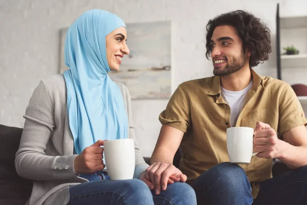 Pasangan Muslim Tersenyum Berpegangan Tangan Dan Memegang Cangkir Kopi Rumah — Stok Foto