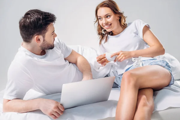 young couple with laptop resting on bed together isolated on grey