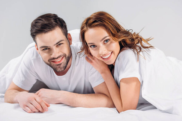 portrait of cheerful young couple in love looking at camera wile lying on bed together isolated on grey
