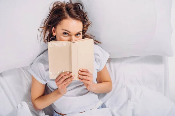 隐藏在床上的妇女覆盖面与书和看相机的看法 — 图库照片