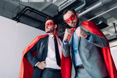 low angle view of handsome super businessmen in masks and capes showing fists in office clipart