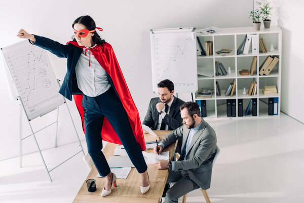 super businesswoman in mask and cape standing on table with hand up in office 