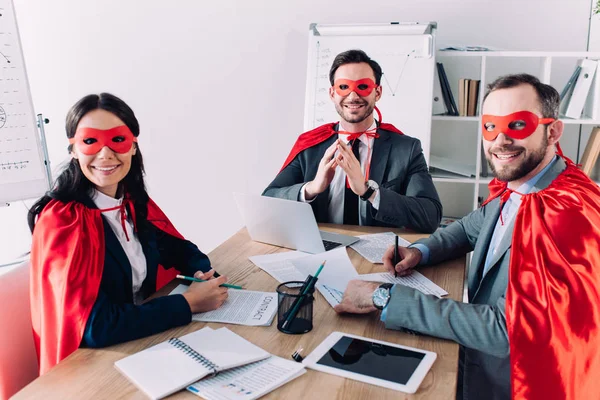 smiling super businesspeople in masks and capes sitting at table in office