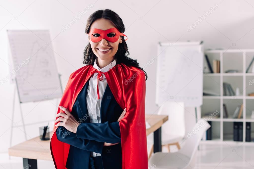attractive super businesswoman in cape and mask standing with crossed arms in office