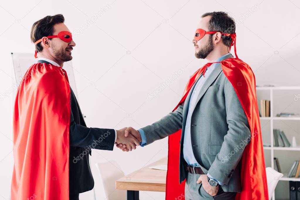 side view of handsome super businessmen in masks and capes shaking hands in office