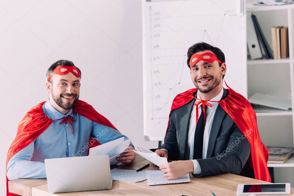 smiling handsome super businessmen in masks and capes looking at camera in office