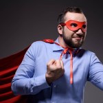 Uśmiechający się przystojny biznesmen super w maskę i pelerynę Wyświetlono pięści na czarnym tle