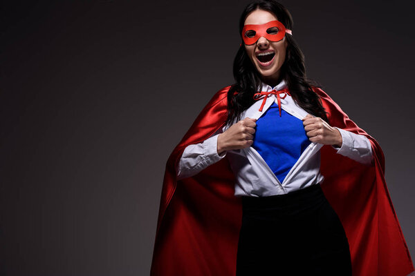 laughing attractive super businesswoman in red cape and mask showing blue shirt isolated on black