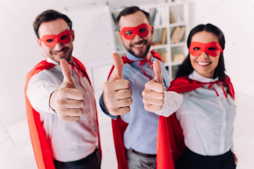 super businesspeople in masks and capes showing thumbs up in office
