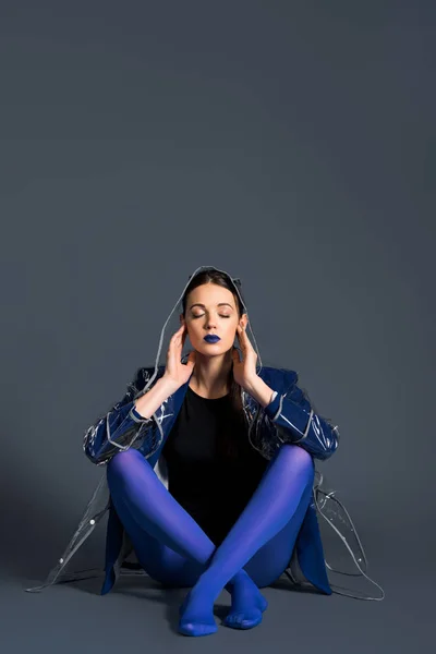 Girl wearing transparent raincoat and blue pantyhose sitting on dark background