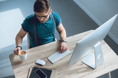 Genç freelancer masaüstü bilgisayarla çalışırken kahve içme havai görünümü