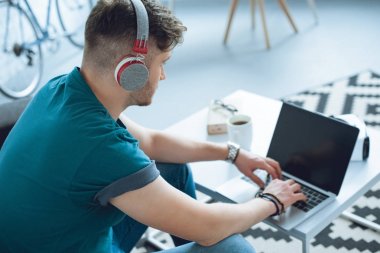 Kulaklıklı genç adam evde dizüstü bilgisayar kullanıyor. 