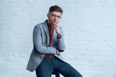 Handsome young man wearing suit and glasses sitting in front of brick wall clipart