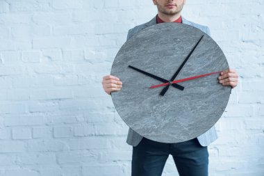 Young man holding big clock in front of him while standing by brick wall clipart